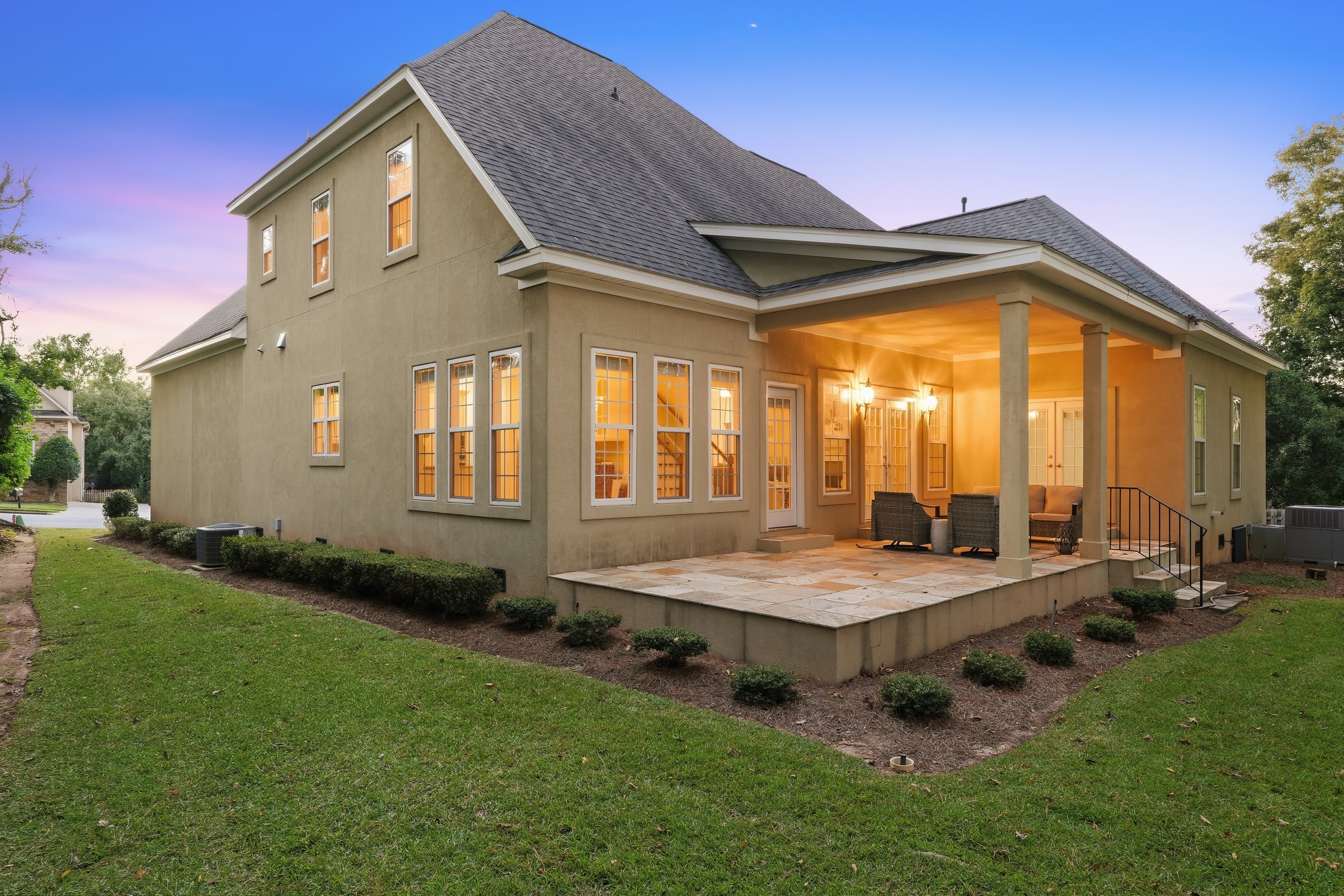 Back of a stucco home at twilight with a lit covered patio and stone terrace