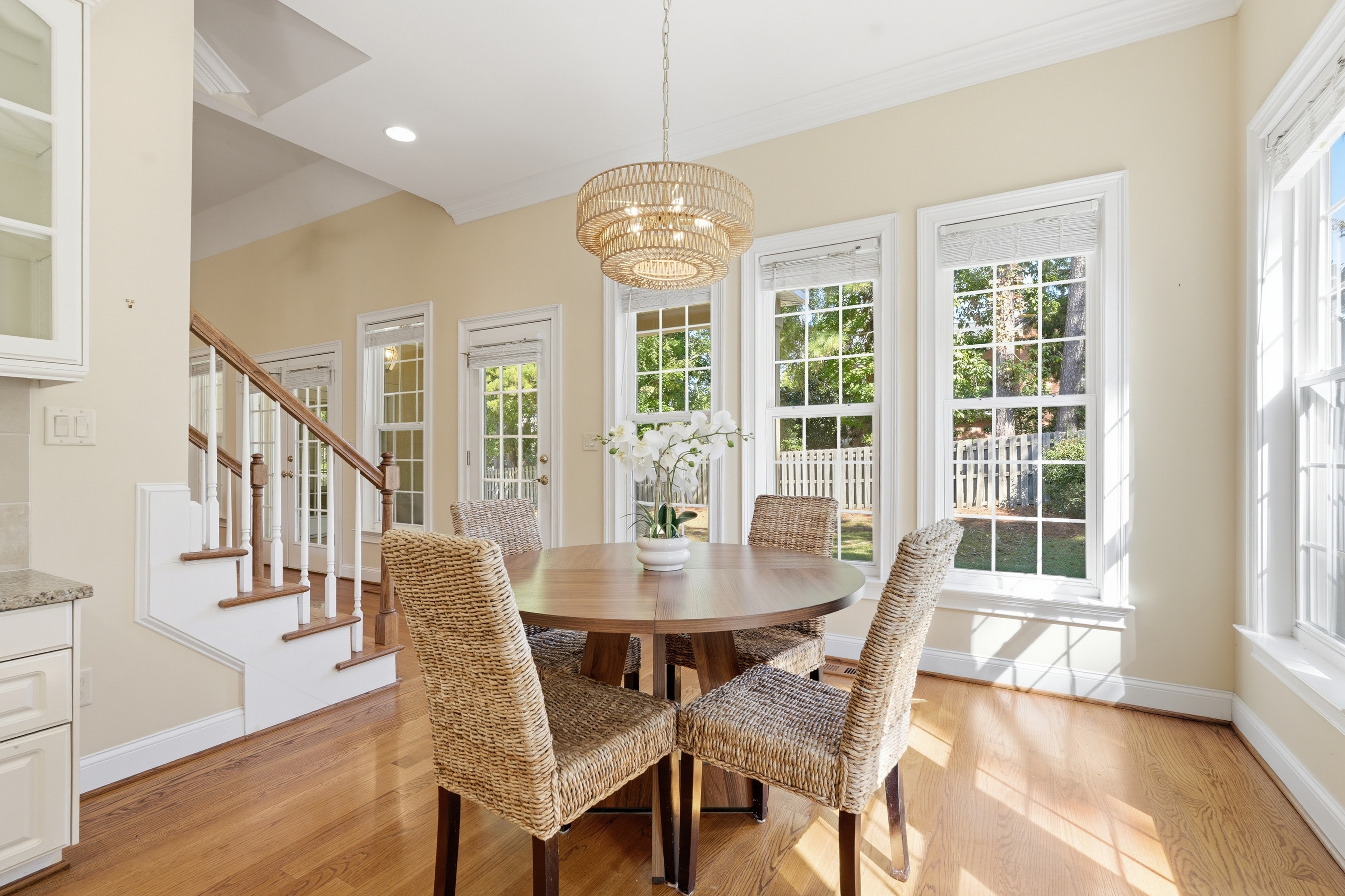 Sunlit breakfast nook with a woven chandelier, round wood table, and tall windows